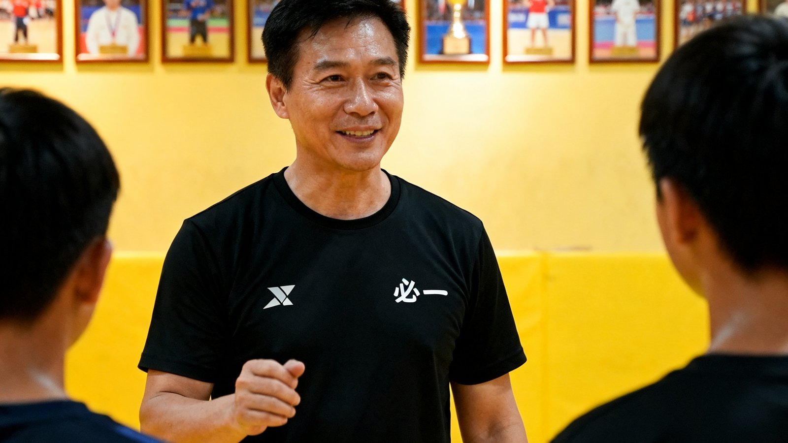 张明辉总教练身穿黑色运动T恤在训练场上指导学员，表情专注而温和，背景是必一运动训练馆的荣誉墙，挂满了学员比赛获奖照片
